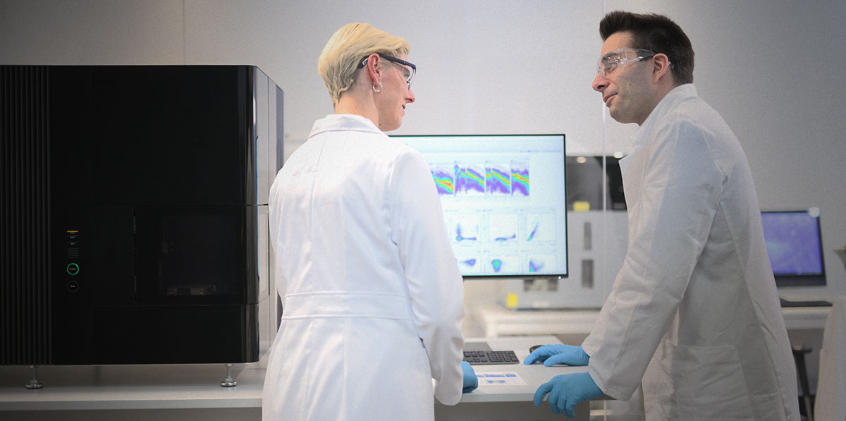 Image of two researchers in a lab engaged in a conversation with an ID7000 cell analyzer and display screen with flow cytometry data